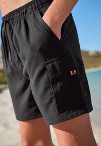 Sorte badebukser med elastisk linning, sidelommer og en cargolomme med et lille pink logo-detalje. Glat tekstur.