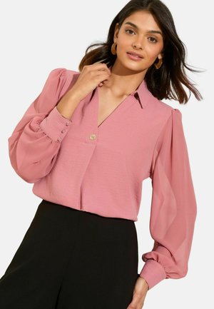 Donna che indossa una blusa rosa con maniche trasparenti a sbuffo e pantaloni neri, mentre si tocca il colletto, con capelli scuri di lunghezza media e orecchini a cerchio.