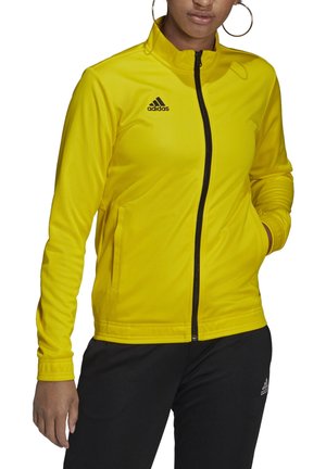 Frau trägt eine knallgelbe Adidas-Zip-Up-Jacke mit schwarzem Adidas-Logo, kombiniert mit schwarzen Adidas-Hosen, Hände in den Jackentaschen.