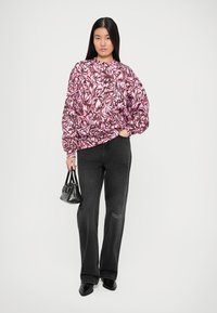 Kvinne iført en oversized genser med rosa og svart mønster, svarte flare-jeans, svarte spisse sko, og holder en liten svart håndveske.