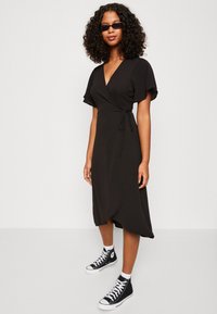 Robe noire portefeuille avec des manches courtes à volants, longueur genou et un lien à la taille, portée avec des baskets montantes noires.