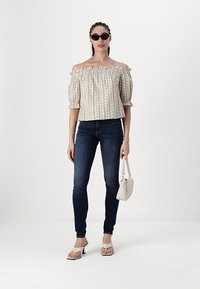 Off-shoulder blomstret topp i beige med puffede ermer, kombinert med mørke, tettsittende jeans. Modellen har en liten hvit håndveske og solbriller.