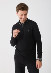 Svart långärmad polotröja med dragkedja i kragen. Har grå kant runt kragen, broderad logotyp på bröstet och en figurnära silhuett.