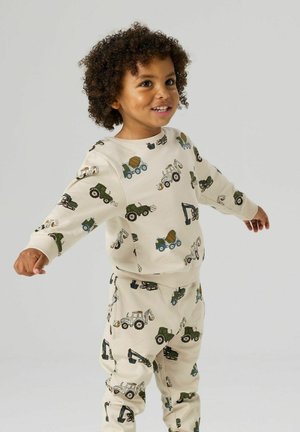 Enfant souriant aux cheveux bouclés portant une tenue crème assortie imprimée de divers engins de chantier, debout les bras étendus.