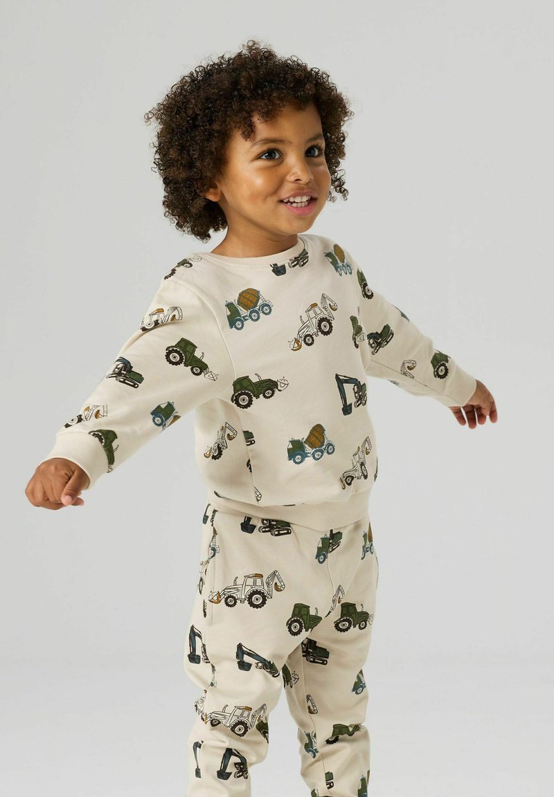 Smilende barn med krøllet hår, iført matchende cremefarvet outfit med print af forskellige byggepladskøretøjer, stående med armene udstrakt.