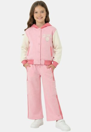 Fille souriante, portant une veste à capuche rose et crème avec le texte « Lee Cooper », un pantalon rose assorti avec des poches latérales, et des baskets blanches.