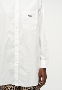 Λευκό πουκάμισο με κουμπιά και μακριά μανίκια, με τσέπη στο στήθος κεντημένη με τη φράση "Moschino Jeans", φορεμένο με παντελόνι σε λεοπάρ μοτίβο.