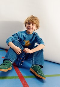 Sweatshirt bleu avec un graphique de chien brun, bouteille d'eau bleue avec un poisson de dessin animé, baskets vertes avec des accents blancs, et jeans bleus.
