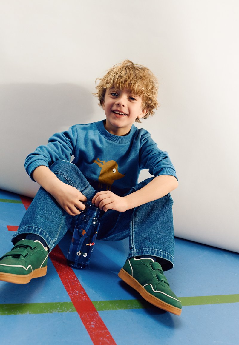 Sweatshirt bleu avec un graphique de chien brun, bouteille d'eau bleue avec un poisson de dessin animé, baskets vertes avec des accents blancs, et jeans bleus.