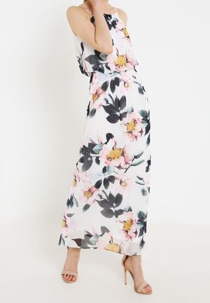 Kobieta ubrana w białą sukienkę maxi bez rękawów z dużym różowym i żółtym kwiatowym wzorem oraz beżowe sandały na wysokim obcasie, pozująca na białym tle.
