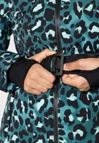 Χέρια που στερεώνουν μια μαύρη αγκράφα ζώνης πάνω από ένα ζακέτα με φερμουάρ σε teal και μαύρο σχέδιο που διαθέτει τρύπες για τους αντίχειρες στα μανίκια.