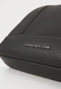 Musta teksturoitu nahkalaukku, jossa on sileä pinta, sisältäen hopean väriset "TOMMY HILFIGER" -brändäykset sivussa ja hienovaraiset ompeluyksityiskohdat.