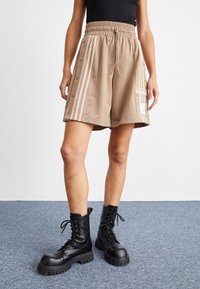 Beige sportshorts med sidoknappar, tre vita ränder och en Adidas-logopatch, som är ihop med svarta snörade kängor.