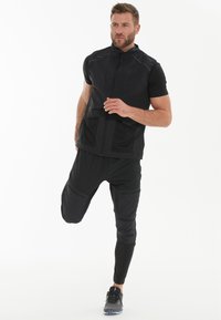 Svart atletisk väst med dragkedja framtill, i kombination med åtsittande svarta byxor med cargofickor och åtsittande muddar. Texturerad tyg för rörlighet.