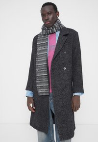 Hall hallabardiga kahe rinnaga kasukas kraega, millel on musta ja halli triibuline sall, roosa T-särgi ja siniste teksade kohal.