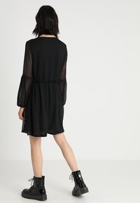 Robe noire jusqu'aux genoux avec manches longues en voile transparent, taille froncée et texture lisse. Elle présente une jupe évasée et est portée avec des bottines noires.