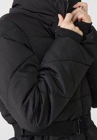 Hands zipping up a black padded jacket with a high collar and a wide belt at the waist, shown from shoulder to mid-torso.