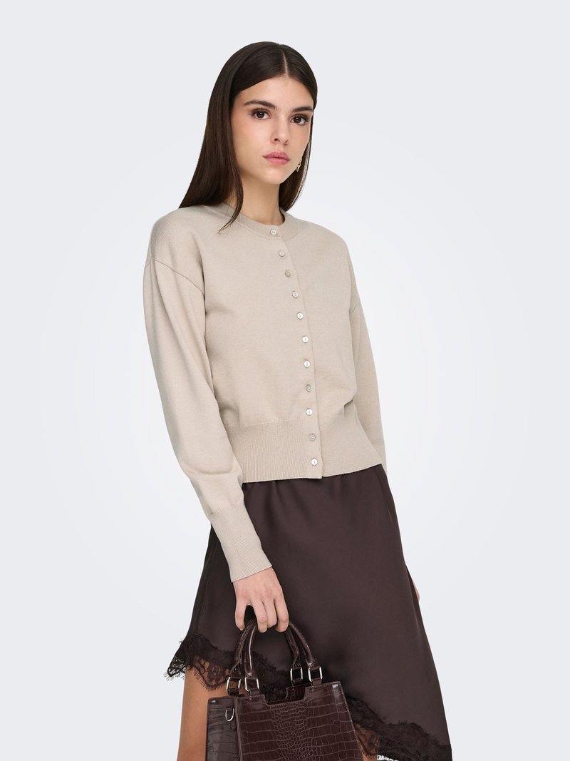 Cardigan court beige avec encolure ronde et boutons en perles, associé à une jupe marron foncé ornée de dentelle. Le modèle tient un sac à main marron foncé.