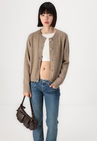 Cardigan de lã bege com um decote redondo, fecho de botão e punhos canelados, combinado com jeans de denim azuis e uma mala de couro castanha.