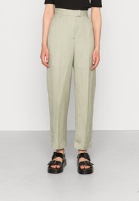 Jasnozielone spodnie z wysokim stanem o luźnym kroju, z przodu z zaszewkami i gładką strukturą, zestawione z czarnymi sandałami na platformie.