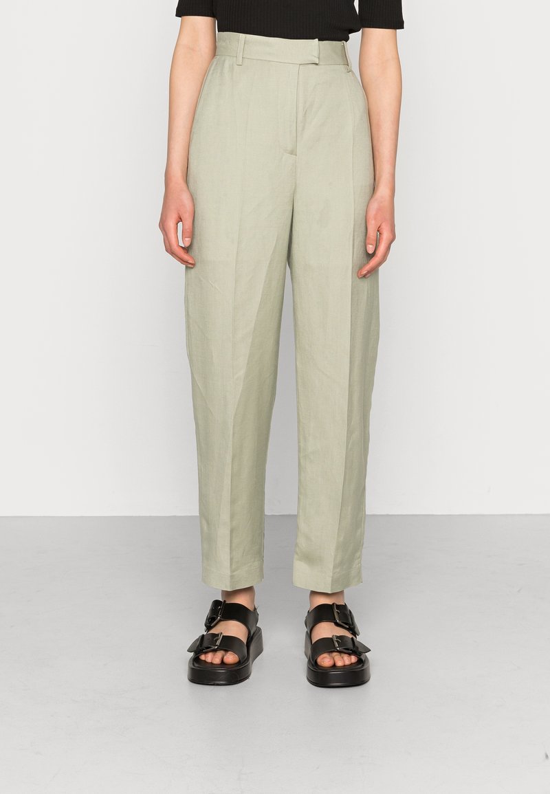 Jasnozielone spodnie z wysokim stanem o luźnym kroju, z przodu z zaszewkami i gładką strukturą, zestawione z czarnymi sandałami na platformie.