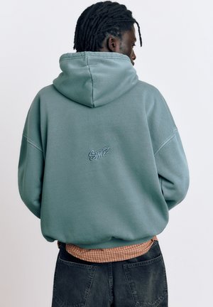 Osoba s dredmi v tyrkysovej mikine s kapucňou s nápisom "Swift" na chrbte, v oranžovej kockovanej košeli a čiernych džínsoch, otočená chrbtom.