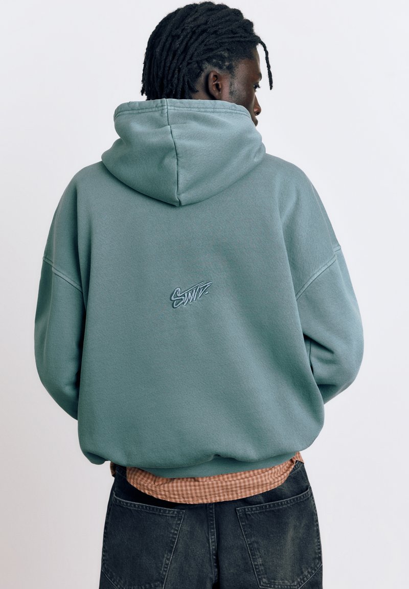 Oseba z dreadlocksi, oblečena v zeleno-modro kapucar z napisom "Swift" na hrbtu, oranžno karo srajco in črne kavbojke, obrnjena stran.