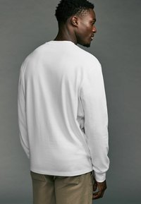 Witte lange mouwen shirt van katoen, met een ronde hals en een ontspannen fit. Het model draagt khaki broeken en staat van achteren geposeerd.