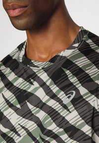 Zelené a černé vzorované sportovní tričko s kulatým výstřihem, které má strukturovanou tkaninu a malé logo na prsou.