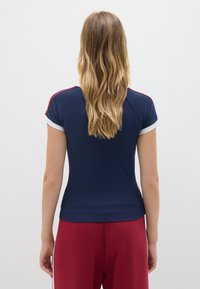 T-shirt blu navy con maniche corte bianche, caratterizzato da accenti rossi sulle spalle. Design aderente, texture liscia e scollatura a girocollo.