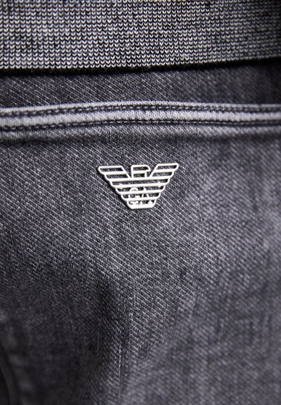 Jeans tkanina u tamnosivoj boji, sa srebrnim metalnim logotipom stilizovanih krila sa brend emblema, postavljenim na području džepa.