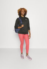 Fekete kapucnis pulóver, rózsaszín leggings és szürke sportcipő. A modell egy kék hátizsákot tart. A háttér egyszínű fehér.