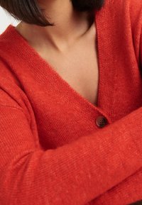 Červený vlněný cardigan s hlubokým výstřihem do V, žebrovanými manžetami a jedním hnědým zapínáním na knoflík. Měkká textura a mírně volný střih.