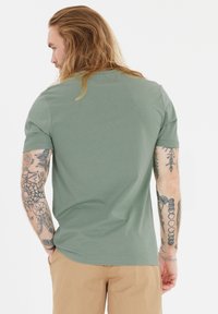 Lyhythihainen, oliivinvihreä t-paita, joka on valmistettu pehmeästä kankaasta ja jossa on pyöreä kaula-aukko. Käsiin näkyvät tatuoinnit tuovat yksityiskohtia ja tekstuuria.