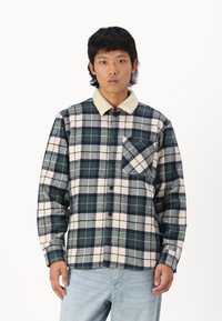 Chemise à carreaux en flanelle bleu marine, vert et crème. Dotée d'un col en sherpa crème doux et d'une poche sur la poitrine avec un motif diagonal. Fermeture par boutons.