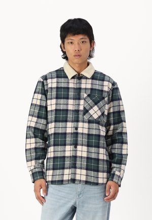 Plaid shirt in navy, green, and cream flannel. Features a soft, cream sherpa collar and a chest pocket with diagonal pattern. Button closure.