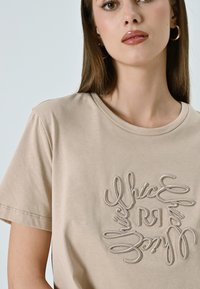 Beige katoenen t-shirt met een ronde hals en korte mouwen, voorzien van ingewikkelde witte borduurwerk in een cirkelvormig patroon aan de voorkant.