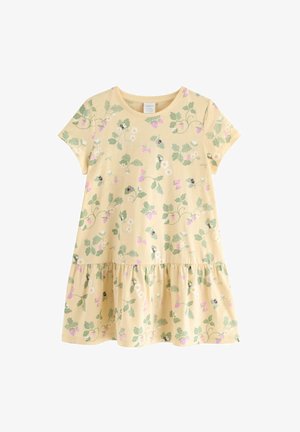 Smilende ung pige i gul blomsterkjole og matchende leggings, iført hvide sneakers, stående mod en enkel lys baggrund.