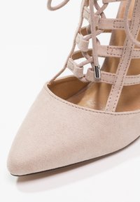 Chaussure en daim beige à bout pointu avec un design de laçage en croix et un accent en métal argenté près des lacets, révélant une partie du pied.
