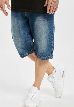 Denim-Shorts in heller Blaufärbung, knielang mit lockerer Passform, kombiniert mit weißen Sportsneakern mit strukturierter Oberfläche und Luftsohle.