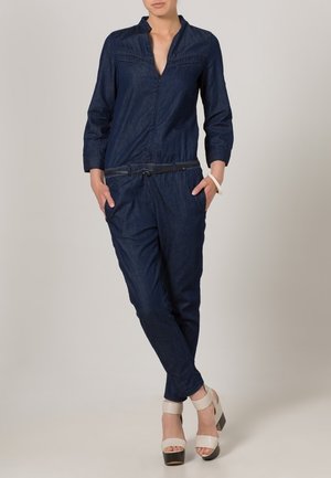 Mono de mezclilla azul marino con cuello camisa, mangas largas y cintura ceñida. Presenta bolsillos frontales y un ligero acento de cinturón. Tacones gruesos.