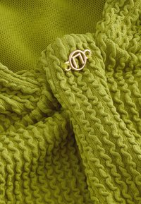 Tissu tricoté vert avec une texture côtelée et un détail de boucle circulaire dorée. Design doux et flexible avec un motif ondulé continu.