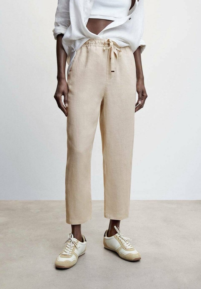 Pantalons en lin beige clair à coupe décontractée, taille élastique et détail de lien. Associés à des baskets de sport blanches et beiges.