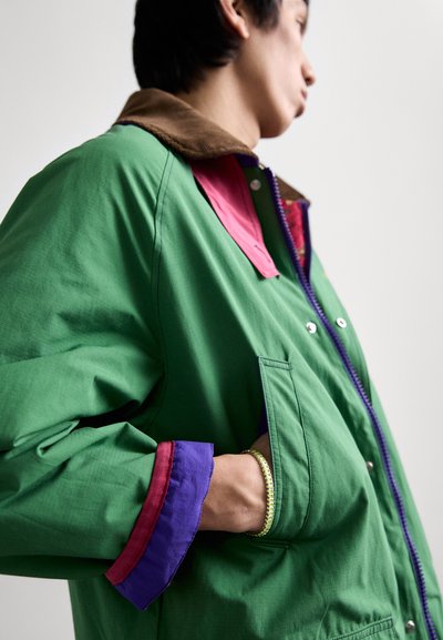 Cette veste colorée présente un extérieur vert, un col en velours côtelé marron et des poignets violets et rouges. Elle est dotée d'une fermeture éclair et de poches latérales.