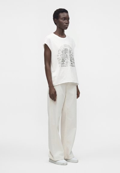 Femme debout portant un t-shirt graphique blanc sans manches avec texte, un pantalon blanc à jambes larges et des baskets blanches sur fond uni.