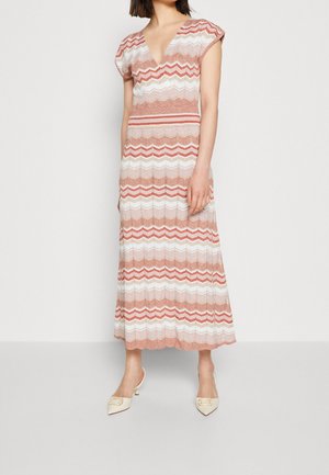 Kobieta w sukience bez rękawów, z dekoltem w szpic, sięgającej kostek, w różowe, białe i czerwone zygzakowate paski, w towarzystwie białych szpiczastych butów na obcasie.