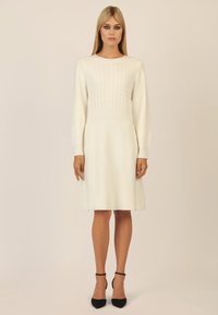 Weißes Strickkleid mit strukturiertem Zopfmuster am Oberteil, langen Ärmeln und ausgestellt Rock. Begleitet von schwarzen Absätzen mit Knöchelriemen.