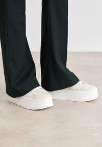 Białe sneakersy na platformie z teksturowaną podeszwą i białymi sznurówkami. Noszone z czarnymi spodniami, prezentując czysty i nowoczesny design.