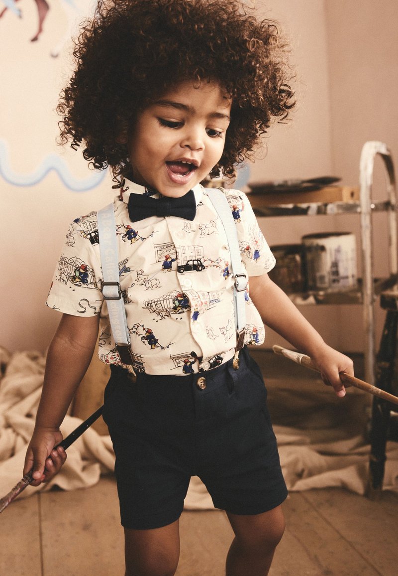 Jeune enfant aux cheveux bouclés portant un nœud papillon, une chemise imprimée et des bretelles, tenant des pinceaux dans un atelier d'art.