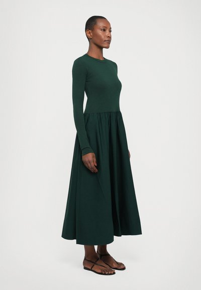 Long-sleeve, dark green dress made of cotton, fitted at the bodice with a flared, ankle-length skirt. Worn with black sandals.